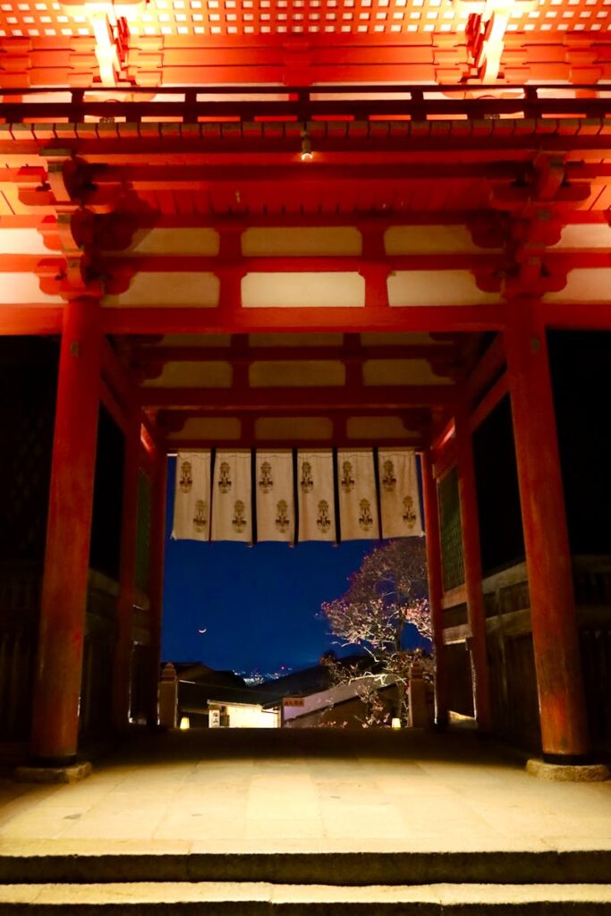 Night view of a traditional Japanese temple gate glowing in warm light, under a crescent moon in Kyoto, Japan.